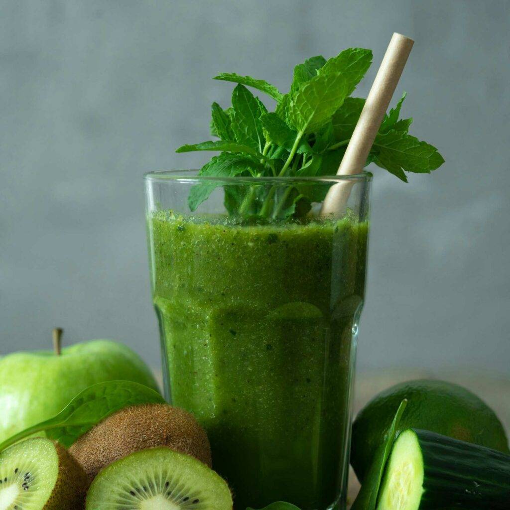 Ein Bild von einem Glas gefüllt mit grünem Smoothie, drum herum liegen grüne Früchte und Gemüse. Als Symbol von Gesundheit.