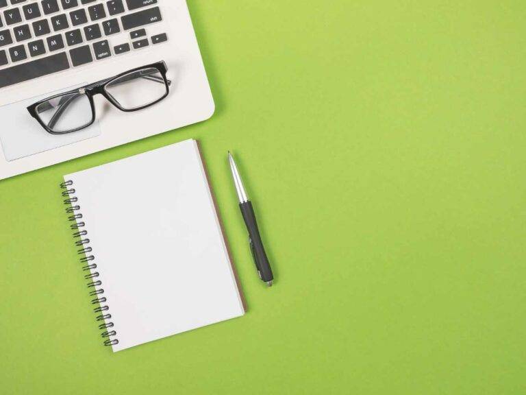 Das Bild zeigt einen aufgeklappten Laptop auf einem grünen Untergrund. Neben ihm liegen sehr ordentlich ausgerichtet ein weißer leerer Block, ein Kugelschreiber und eine Brille. Es symbolisiert Ordnung und Management.