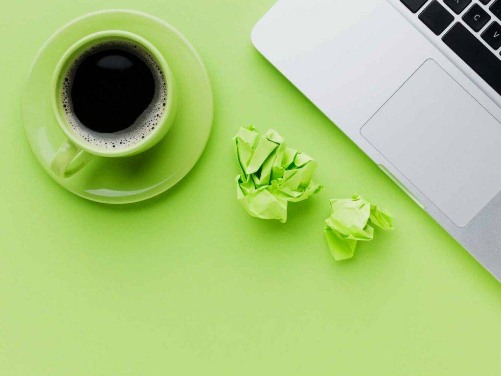 Auf dem Bild sieht man einen gefüllte Kaffeetasse, die Ecke von einem Laptop und zwei geknüllte Papierkugeln. Alles auf einem grünen Untergrund. Es symbolisiert einen Arbeitsplatz.