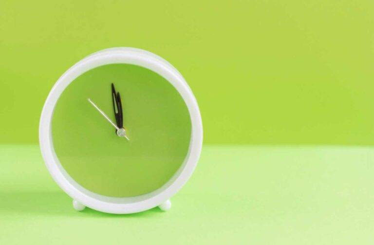 Ein Bild einer weißen Uhr vor einem grünen Hintergrund. Beide Zeiger stehen auf kurz vor 12. Das Bild symbolisiert Zeitmanagement sowie bevorstehende Veränderungen.