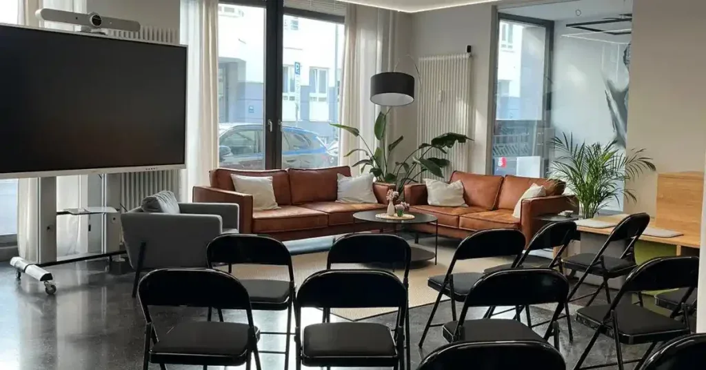 Veranstaltungsfläche GreenlightHUB im modernen Lounge-Design – ideal, um ein Business Event zu planen. Im Vordergrund halbkreisförmig aufgestellte schwarze Klappstühle vor einem großen Bildschirm, im Hintergrund gemütliche Sitzgruppe mit braunen Ledersofas. Perfekter Ort für Meetings, Workshops oder Events wie die Veranstaltung Greenlight Connect.