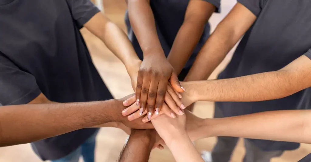 Nahaufnahme, die zeigt, wie mehrere Arme unterschiedlicher Hautfarben in der Mitte aufeinandergelegt werden, als Symbol für Teamwork, Gemeinschaft und Zusammenhalt. Das Bild eignet sich gut zur Veranschaulichung des Tag des Ehrenamts oder anderer ehrenamtlicher Aktivitäten.
