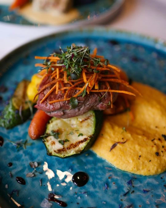 Hochwertig angerichteter Hauptgang mit zartem Fleisch, gegrilltem Gemüse und cremigem Püree auf einem blauen Teller – serviert als Teil eines exklusiven 3-Gänge-Menüs.