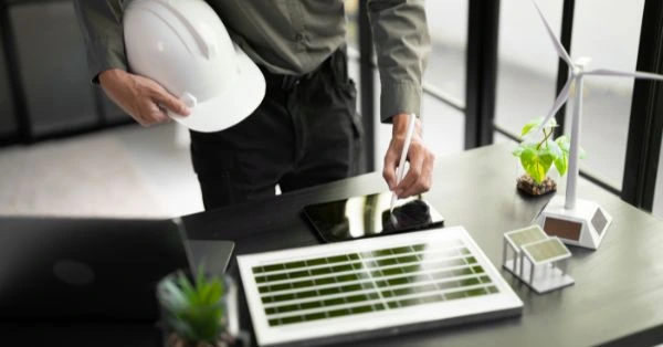 Eine Person hält einen Schutzhelm in der Hand und bedient ein Tablet auf einem Schreibtisch, auf dem ein Solarpanel-Modell, eine kleine Windkraftanlage und Architekturmodelle stehen – Symbol für digitale Planung, Nachhaltigkeit und moderne Ingenieurarbeit.