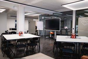 Workshopraum im GreenlightHUB München mit Inselbestuhlung (Gruppentische), Screen, Flipchart und modernem Ambiente.