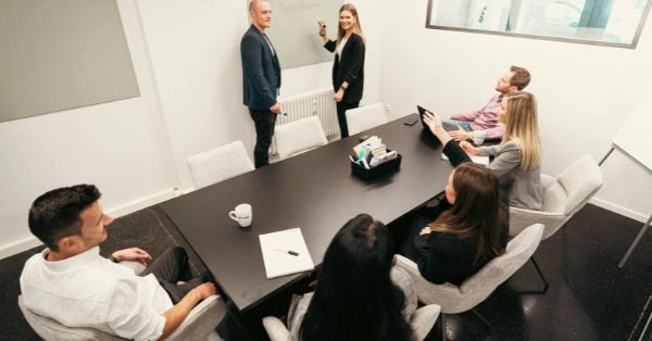 Meeting-Szene im Rahmen von Corporate Events: Mehrere Personen sitzen in einem modernen Konferenzraum an einem Tisch, während zwei Personen vorne präsentieren. Die Aufnahme zeigt eine professionelle Workshop- oder Event-Situation mit Fokus auf Austausch, Zusammenarbeit und Wissenstransfer.