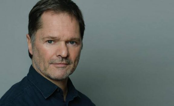 Porträt von Frieder Gamm - Speaker Greenlight Connect 2026 zum Thema KI in der Verhandlungsführung. Porträt eines Mannes mit kurzem dunklem Haar, dunklem Hemd und ernstem Blick vor grauem Hintergrund.